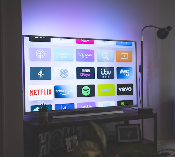 A flat-screen television is displaying a variety of streaming app icons, including Netflix, Prime Video, Spotify, and ITV Hub among others. The TV is placed on a media console with a small potted plant beside it. A tall lamp stands to the right, and a faint, ambient light casts a purple hue on the wall above the TV.